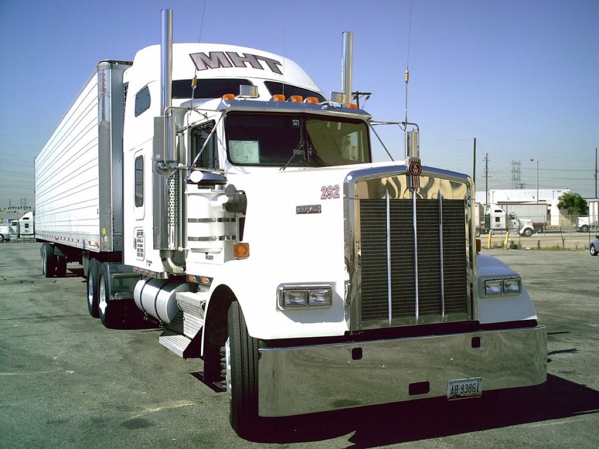 A white Kenworth tractor pulling a dry-van trailer, parked at a truck stop